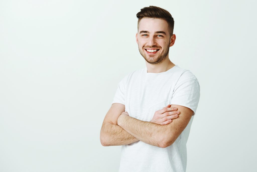 Portrait of pleased confident and accomplished european guy with stylish haircut standing unshaven over gray background crossing hands on chest smiling joyfully and satisfied at camera